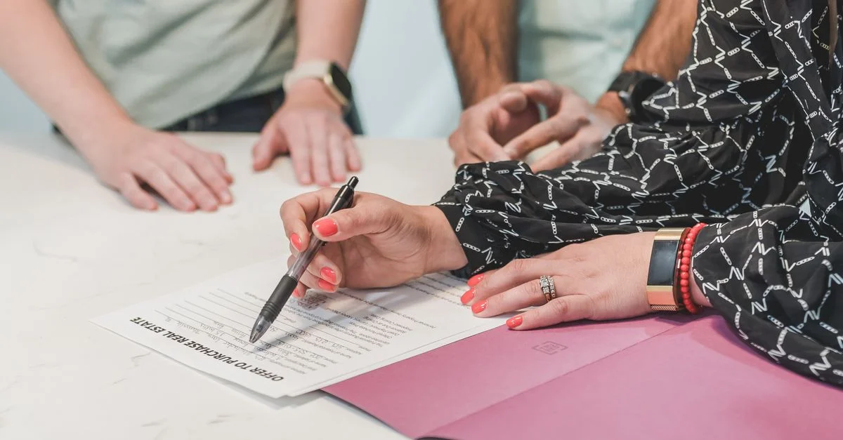 Gros plan sur des mains signant un document immobilier à une table de réunion avec trois personnes.